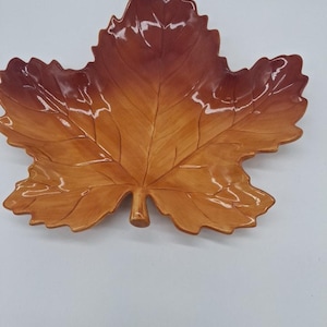 May include: A ceramic dish shaped like a large maple leaf with a glossy finish. The color transitions from a deep reddish-brown at the top edges to a lighter, warm golden-brown near the stem, featuring embossed vein details across the surface.