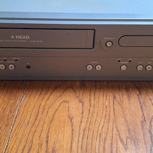 May include: Silver Magnavox DVD/VCR combo player. The unit has a VCR slot labeled "4 HEAD" and a DVD tray. It features multiple control buttons and the DVD logo. The device is on a wooden surface.