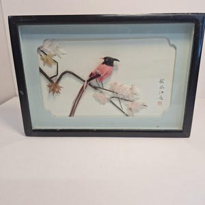 May include: A framed artwork featuring a pink and black bird perched on a branch with delicate white and yellow flowers. The bird has a long tail and is set against a light blue background. The frame is black.