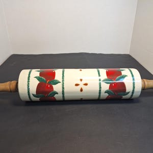 May include: A ceramic rolling pin with a cream-colored base, adorned with red apple designs and green leaves. The handles are made of wood. This kitchen tool is designed for rolling out dough.