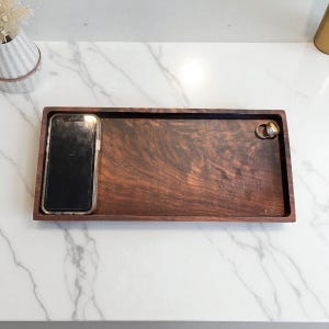 May include: A rectangular wooden tray with a dark brown finish. A smartphone in a clear case rests in a recessed area. Two rings are on the right side. The tray has the engraved text "W A T E R".