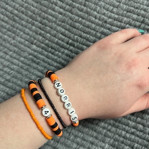Peut inclure: Quatre bracelets empilés sur un poignet. Le bracelet du haut est rayé noir et orange avec des perles blanches qui épellent le nom "NORRIS". Le deuxième bracelet est noir avec des perles blanches qui épellent le nombre "4". Le troisième bracelet est rayé noir et orange. Le bracelet du bas est orange.