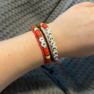 Peut inclure: Un ensemble de quatre bracelets avec des perles rouges, noires et jaunes. Un bracelet porte le nom "Leclerc" écrit en lettres blanches sur des perles noires. Un autre bracelet porte le numéro "16" écrit en lettres blanches sur des perles rouges.