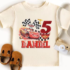A white t-shirt with a red and black design featuring a race car and the number 5, along with a pair of brown baby shoes and a white camera.