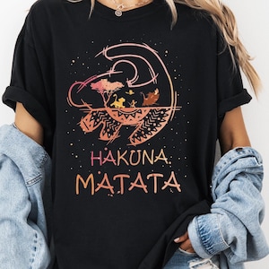 May include: Black t-shirt featuring a colorful graphic of a lion's head with the text "HAKUNA MATATA" in orange. The design includes a savanna scene. The shirt is a comfortable, casual style.