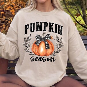 May include: A beige crewneck sweatshirt featuring a graphic of an orange pumpkin with a black and white polka dot bow, surrounded by black leaf illustrations and the text PUMPKIN SEASON in a bold, distressed black font across the chest.