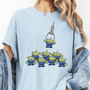 A woman wearing a light blue t-shirt with a graphic of a group of alien characters being lifted by a crane.