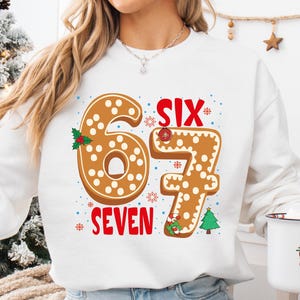 May include: White sweatshirt with a gingerbread-style "67" design and the word "SEVEN" in red. The design includes holly and a Christmas tree. A white mug with a Santa Claus illustration is held in the hand.