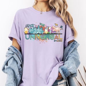 Może przedstawiać: Jasnofioletowa koszulka z kolorową grafiką postaci i atrakcji Universal Studios. Tekst "UNIVERSAL STUDIOS" jest wyświetlany w tęczowym gradiencie. Koszulka jest wykonana z miękkiego materiału.
