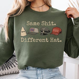 May include: Olive green sweatshirt with the text "Same Shit. Different Hat." and illustrations of a KKK hood, a Confederate flag hat, a Nazi hat, and a "Make America Great Again" hat. The sweatshirt is worn by a person.