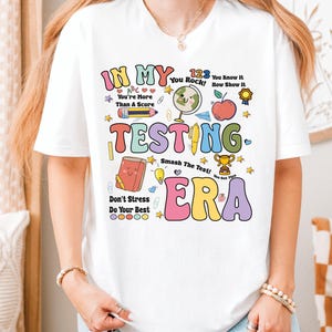 May include: White t-shirt with a colorful graphic design that says "In My Testing Era". The graphic includes illustrations of school supplies, hearts, stars, and a trophy.