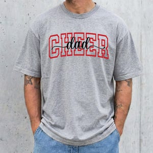 Op de afbeelding: Een grijs t-shirt met het woord "CHEER" in rode blokletters en het woord "dad" in zwart cursief. Het shirt heeft een casual stijl en wordt gecombineerd met een blauwe jeans.