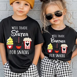 Two children wearing matching black t-shirts with text and illustrations, standing against a white brick wall.