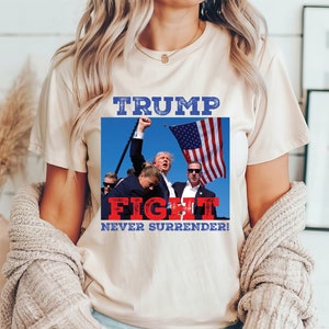 May include: A white t-shirt with a graphic print of a group of people with an American flag. The text on the shirt reads "TRUMP FIGHT NEVER SURRENDER!"