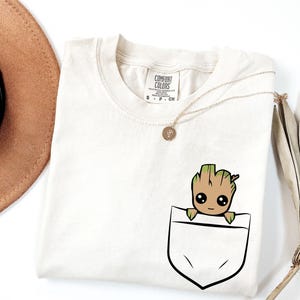 May include: Cream-coloured t-shirt with a pocket design featuring a cartoon Groot character. The shirt has a crew neck and short sleeves. A gold necklace with a pendant is draped across the shirt. The shirt is labelled "Comfort Colors".