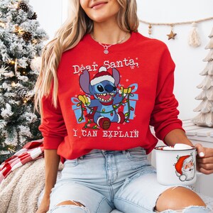 A woman wearing a red sweater with a cartoon character and text, sitting in front of a Christmas tree.