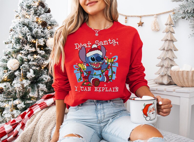 A woman wearing a red sweater with a cartoon character and text, sitting in front of a Christmas tree.