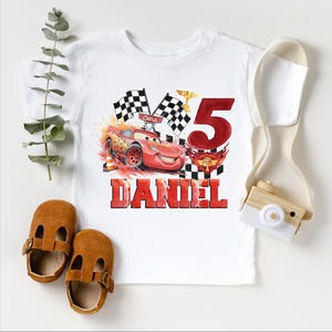 A white t-shirt with a graphic of a red race car and the number 5, along with a pair of brown baby shoes and a camera.