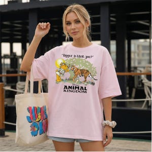 A woman wearing a pink t-shirt with a cartoon tiger and text, standing in front of a building.