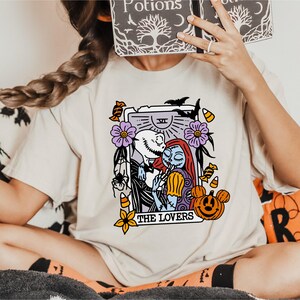 A person is sitting on a couch wearing a white t-shirt with a graphic design featuring characters from the movie "The Jack Skellington".