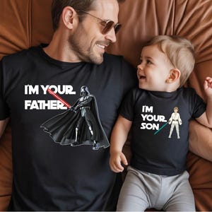 Könnte beinhalten: Schwarze T-Shirts mit Star Wars-Motiven. Eines zeigt Darth Vader mit dem Text "I'M YOUR FATHER". Das andere zeigt Luke Skywalker mit dem Text "I'M YOUR SON".