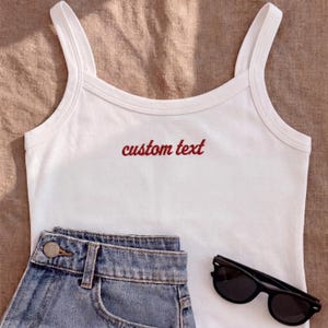 May include: White tank top with thin straps and the embroidered text "custom text" in red. The top is paired with denim shorts and black sunglasses. The items are arranged on a neutral-toned surface.