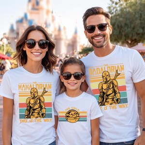 A family of three, consisting of a woman, a man, and a child, is standing together and smiling. They are all wearing white t-shirts with the same design featuring a character from the Star Wars franchise.