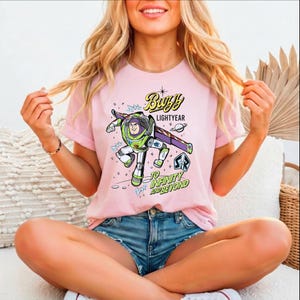 A woman wearing a pink t-shirt with a graphic of Buzz Lightyear from the Toy Story franchise, sitting cross-legged on a couch.