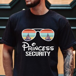 Może przedstawiać: Czarny T-shirt z białym nadrukiem przedstawiającym okulary przeciwsłoneczne z odbitym w szkle zamkiem Disneya. Pod okularami znajduje się napis "Princess Security".