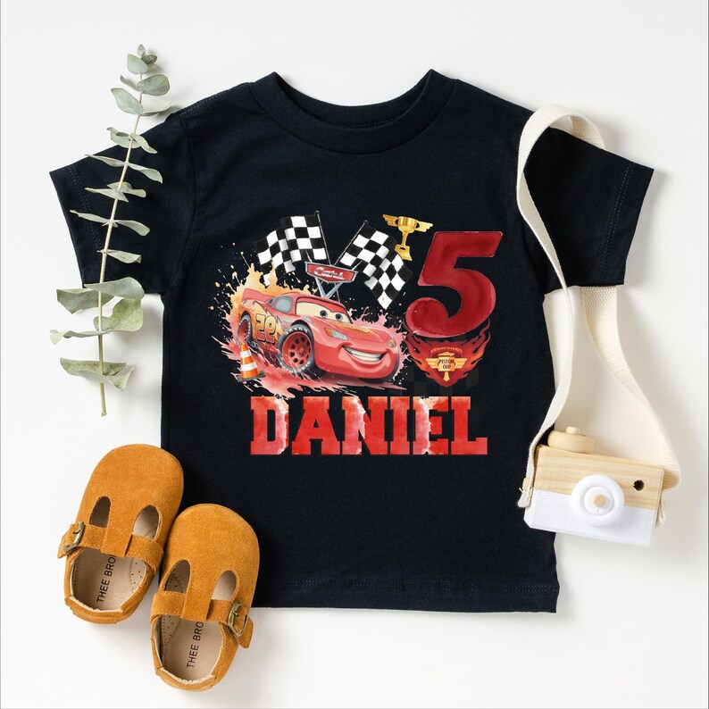 A black t-shirt with a red and white design featuring a race car and the number 5, along with a pair of brown baby shoes and a white camera.