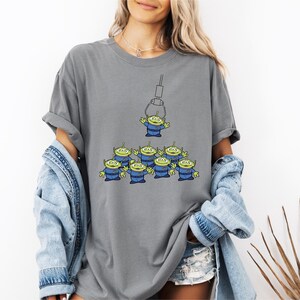 A woman wearing a gray t-shirt with a graphic of a group of alien characters, along with denim clothing and a denim jacket.