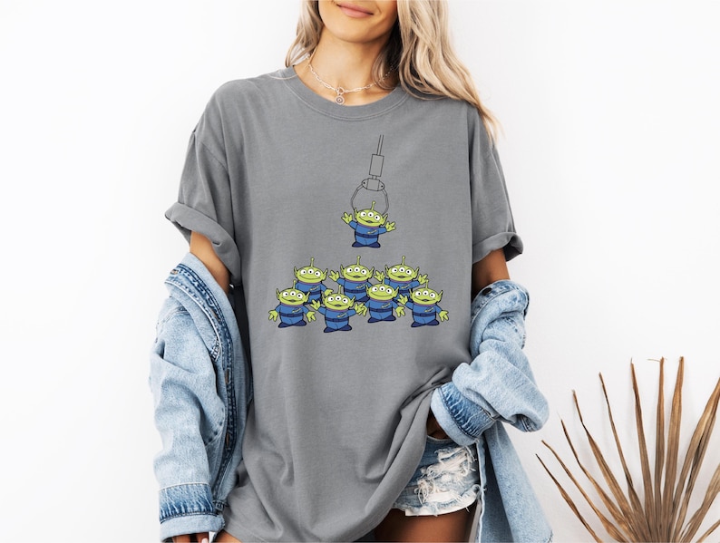 A woman wearing a gray t-shirt with a graphic of a group of alien characters, along with denim clothing and a denim jacket.