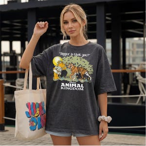 A woman wearing a grey t-shirt with a graphic of a tiger and the text "ANIMAL KINGDOM" is standing in front of a building. She is holding a white tote bag with colorful text on it.