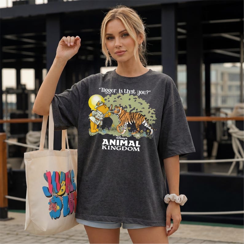 A woman wearing a grey t-shirt with a graphic of a tiger and the text "ANIMAL KINGDOM" is standing in front of a building. She is holding a white tote bag with colorful text on it.