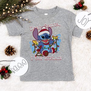A gray t-shirt with a cartoon character and text, surrounded by Christmas decorations such as pine cones, ornaments, and a "JOY" sign.