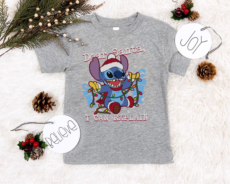A gray t-shirt with a cartoon character and text, surrounded by Christmas decorations such as pine cones, ornaments, and a "JOY" sign.