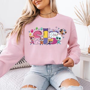 A woman wearing a pink sweatshirt with a colorful graphic design featuring cartoon characters and the text "SUPER KITTES".