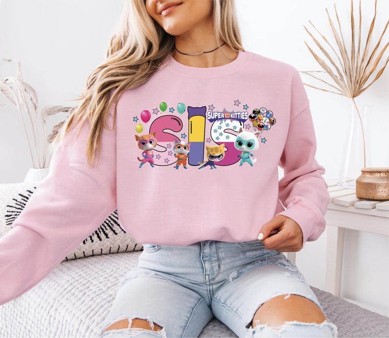 A woman wearing a pink sweatshirt with a colorful graphic design featuring cartoon characters and the text "SUPER KITTES".