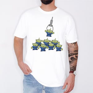 A white t-shirt with a graphic design depicting a group of green alien characters being lifted by a crane.