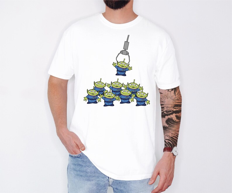 A white t-shirt with a graphic design depicting a group of green alien characters being lifted by a crane.