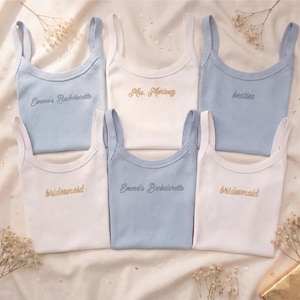 May include: Six tank tops in white and light blue are arranged on a cream-colored surface. Each top has text embroidered on the front in gold or blue thread. The text includes "Emma's Bachelorette", "Mrs. Martinez", "besties", and "bridesmaid".