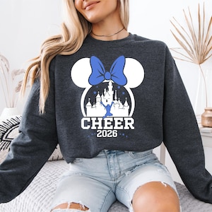 A woman wearing a gray sweatshirt with a graphic design of a castle and a blue bow on it, along with the text "CHEER 2026".