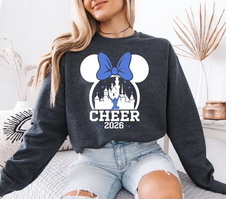 A woman wearing a gray sweatshirt with a graphic design of a castle and a blue bow on it, along with the text "CHEER 2026".