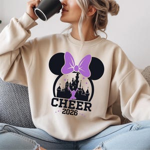 A woman wearing a white sweatshirt with a graphic of a castle and a purple bow, sitting on a couch and drinking from a black mug.