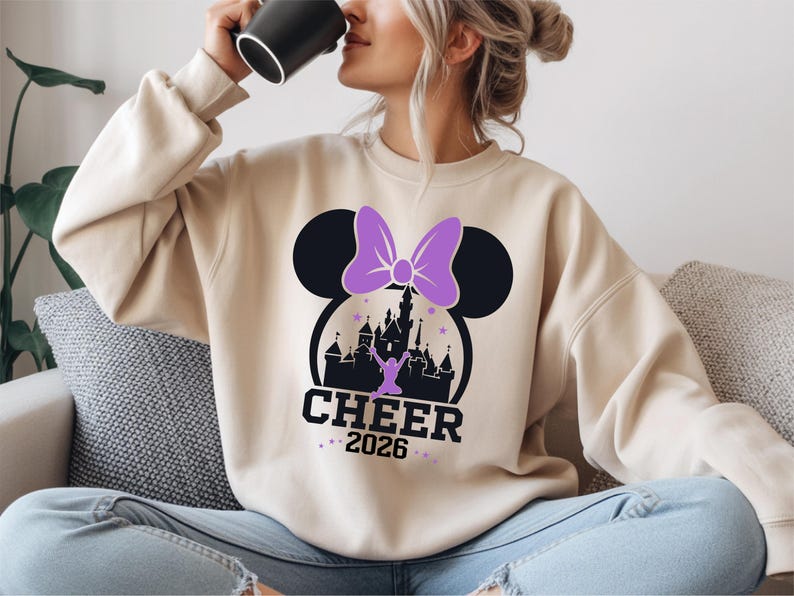 A woman wearing a white sweatshirt with a graphic of a castle and a purple bow, sitting on a couch and drinking from a black mug.