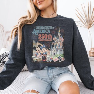 A woman wearing a gray sweatshirt with a graphic design of Disney characters and landmarks, celebrating the United States of America's 250th anniversary.