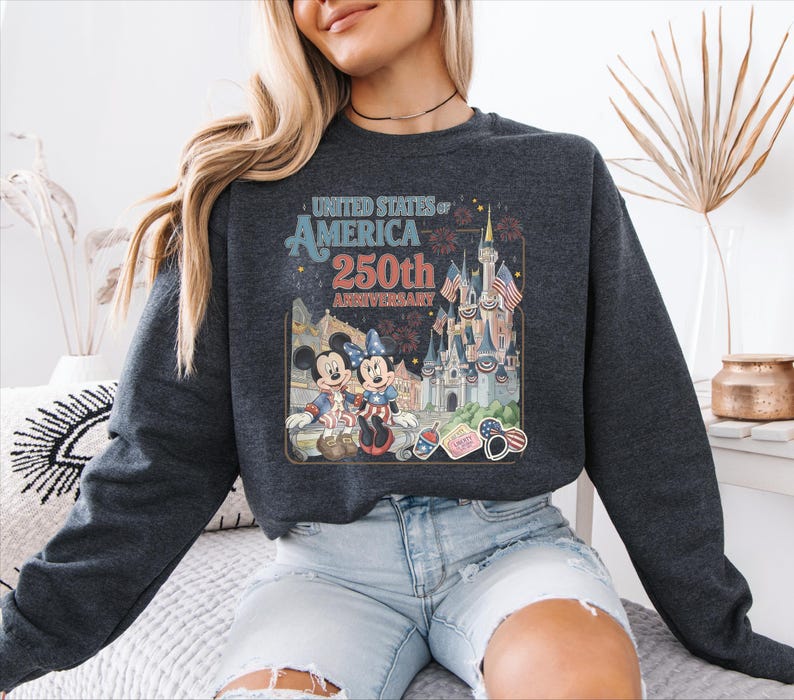 A woman wearing a gray sweatshirt with a graphic design of Disney characters and landmarks, celebrating the United States of America's 250th anniversary.