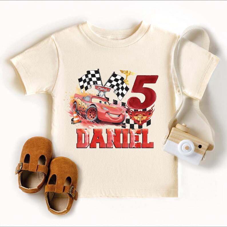 A white t-shirt with a red and black design featuring a race car and the number 5, along with a pair of brown baby shoes and a white camera.