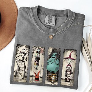 May include: Gray Comfort Colors t-shirt featuring four panels with characters from The Nightmare Before Christmas. The panels include Jack Skellington, Sally, Oogie Boogie, and the Mayor. A silver necklace with a pendant is also visible.