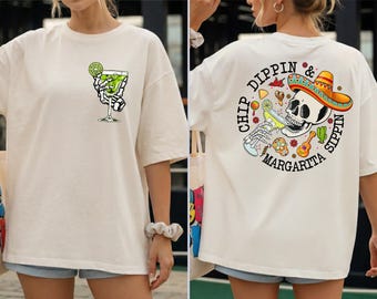 May include: Off-white t-shirt with a skeleton hand holding a margarita glass on the front. The back of the shirt features a skull wearing a sombrero, surrounded by the words "Chip Dippin' & Sippin' Margarita."